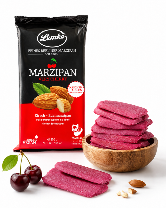 Lemke Very Cherry marzipan package with pink cherry marzipan pieces in a wooden bowl, cherries, and almonds on a white background.