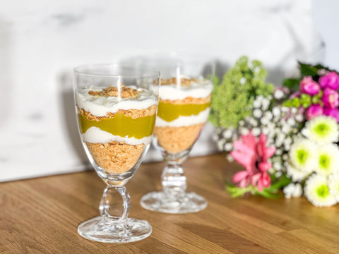 wei Dessertgläser mit Lemke High-Protein Pistazien-Cheesecake – Keksboden, Joghurt und Pistazienmus in Schichten; Blumen im Hintergrund.
