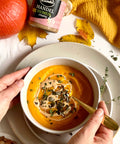 A jar of Lemke Mandel BioMus creamy almond butter next to a bowl of pumpkin soup garnished with seeds and herbs. A person is holding a spoon in the bowl, ready to eat. The setting includes autumn leaves, a bright orange pumpkin, and a cozy yellow fabric.