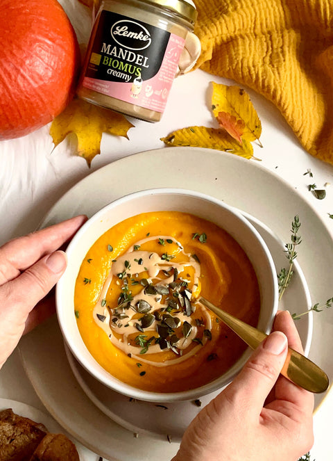 A jar of Lemke Mandel BioMus creamy almond butter next to a bowl of pumpkin soup garnished with seeds and herbs. A person is holding a spoon in the bowl, ready to eat. The setting includes autumn leaves, a bright orange pumpkin, and a cozy yellow fabric.