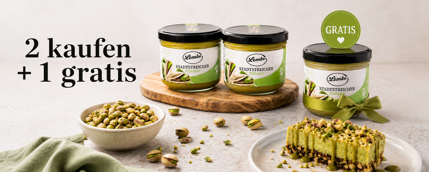 Promotional product image showing three jars of Lemke Stadtstreicher Pistazie pistachio spread on a light background, with two jars on a wooden board and one gift jar with a green ribbon and “Gratis” badge, surrounded by pistachios and a pistachio dessert, plus the headline “2 kaufen + 1 gratis.”